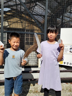 子ども2人がクジャクの翼部分の羽根を持って笑顔でこちらを向いている写真